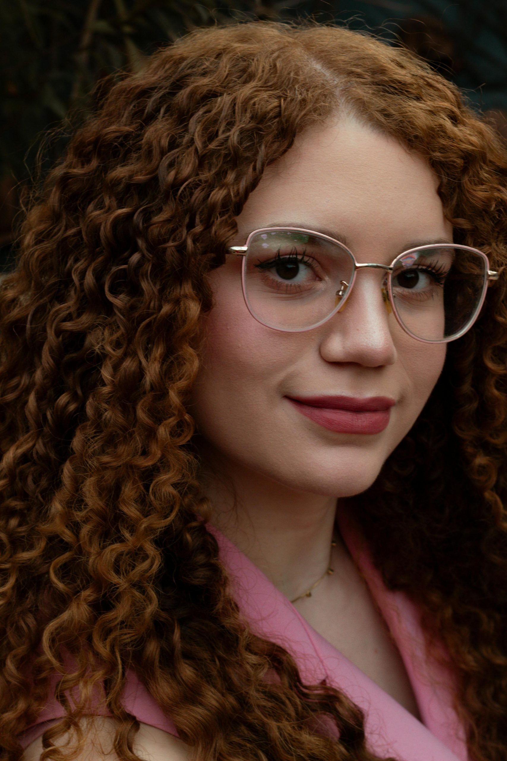 Close-up portrait of a woman with red curly hair and glasses, wearing a pink top.